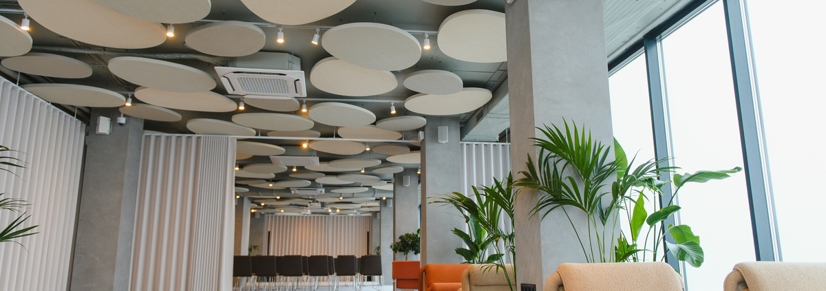 Spacious office lounge with soft seating, tall windows, indoor plants, and round ceiling panels throughout area.