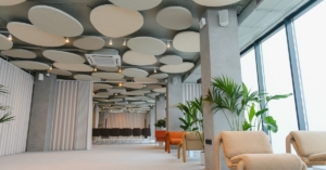 Spacious office lounge with soft seating, tall windows, indoor plants, and round ceiling panels throughout area.