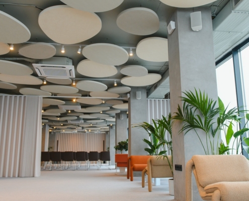 Spacious office lounge with soft seating, tall windows, indoor plants, and round ceiling panels throughout area.