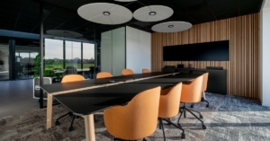 Contemporary meeting space with black table, orange chairs, large screen, and acoustic ceiling panels near glass wall.
