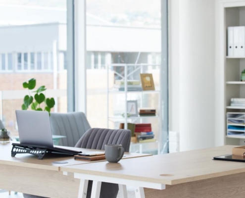 Bright office desk with laptop, notebook, and coffee mug near window, shelves and organized files in background.