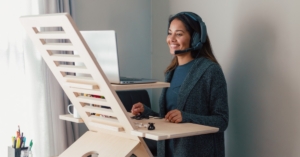 Woman wearing headset smiles while using laptop at standing desk, working from home near window with natural light.