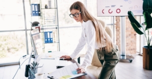 Woman in glasses reviews documents at desk, leaning over computer keyboard in bright office with charts on screen.
