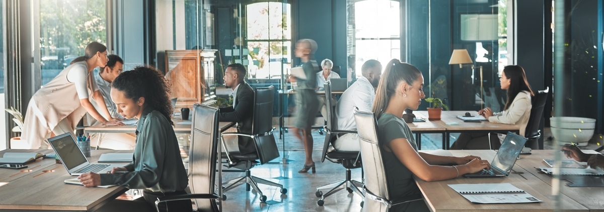 Busy office scene with professionals typing on laptops, meeting and moving through bright open-plan workspace.