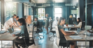 Busy office scene with professionals typing on laptops, meeting and moving through bright open-plan workspace.