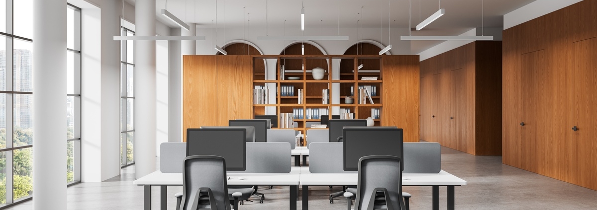 Contemporary workspace with shared desks, computer screens, office chairs, and built-in wooden cabinets and bookshelves.