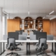 Contemporary workspace with shared desks, computer screens, office chairs, and built-in wooden cabinets and bookshelves.