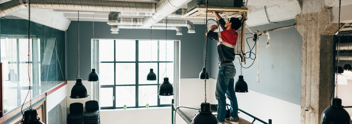 Worker stands on scaffolding installing ceiling equipment in an industrial room with exposed ducts and pendant lights.