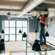 Worker stands on scaffolding installing ceiling equipment in an industrial room with exposed ducts and pendant lights.