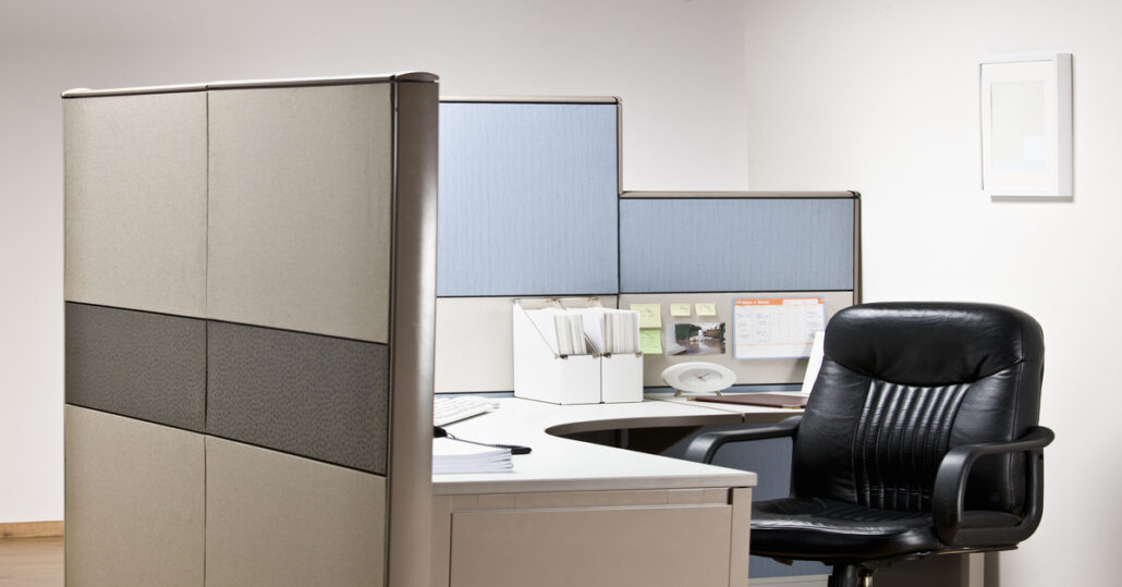 A black leather empty chair waits at tidy desk in cubicle that has blue shelves and white folder organizers.